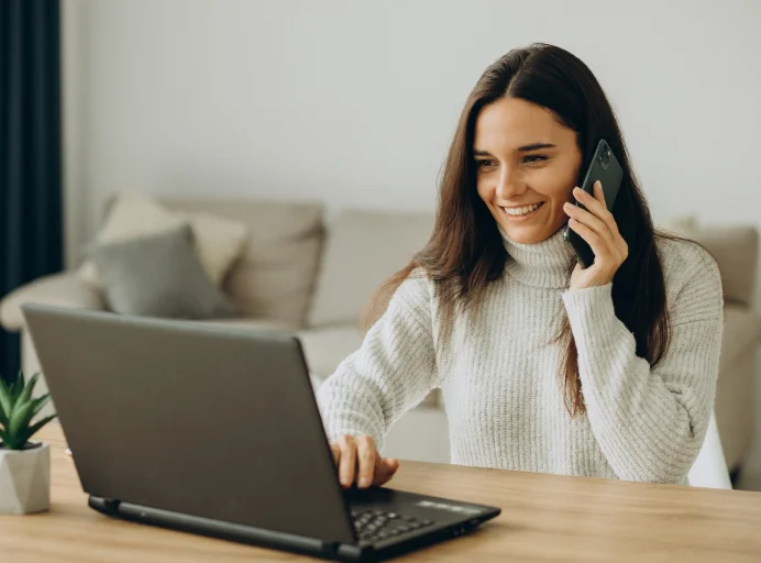 A woman sits at a desk at home smiling while talking on the phone and using a laptop in a comfortable living space. The image represents a working professional exploring an online MBA with flexible learning and remote career support.